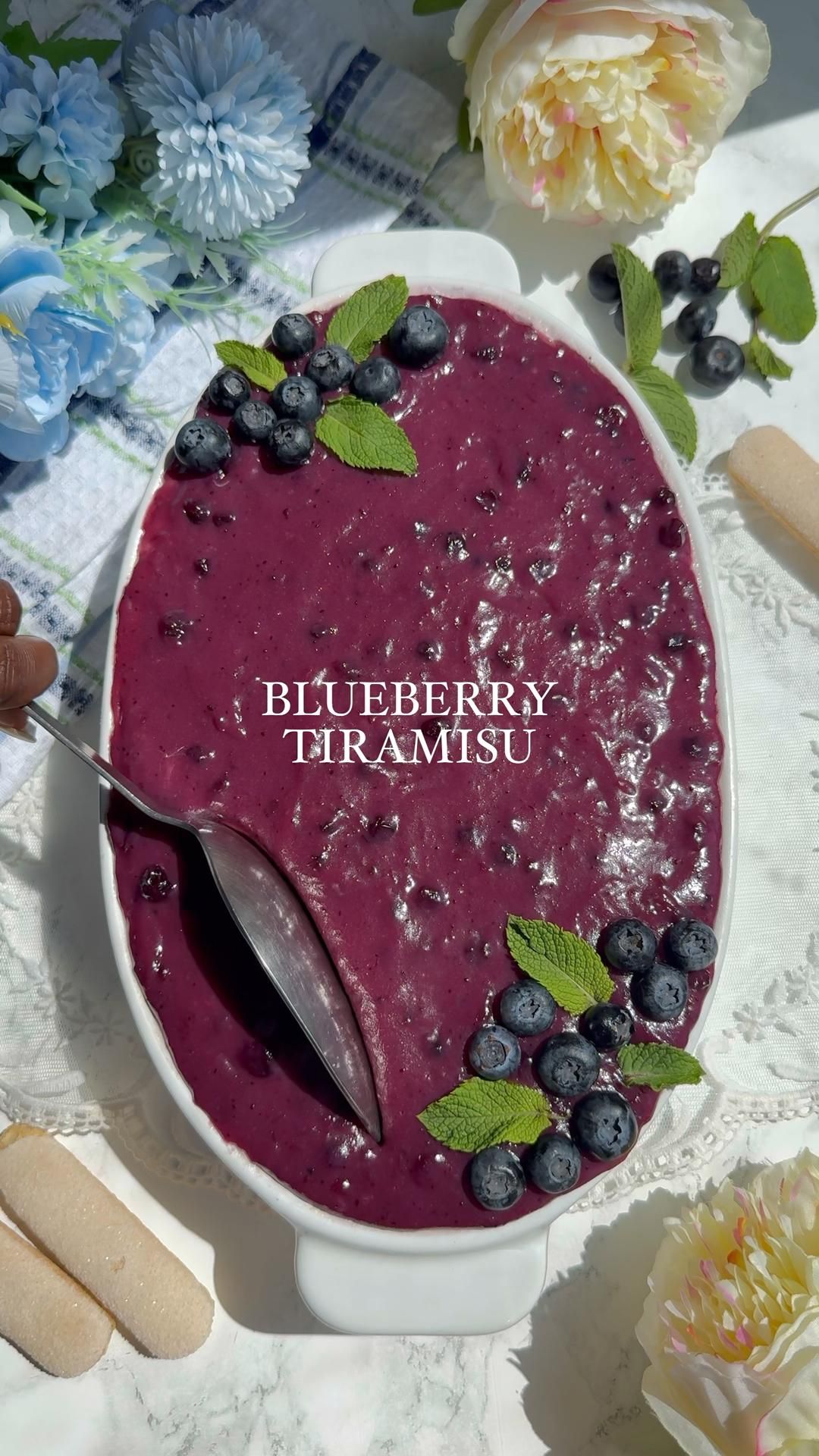 Blueberry tiramisu with fresh blueberries and mint leaves on top, with a serving spoon and ladyfingers on the side.