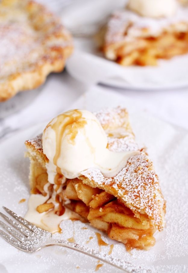 Slice of apple pie topped with melting vanilla ice cream and a drizzle of caramel on a white surface, with a fork beside it.