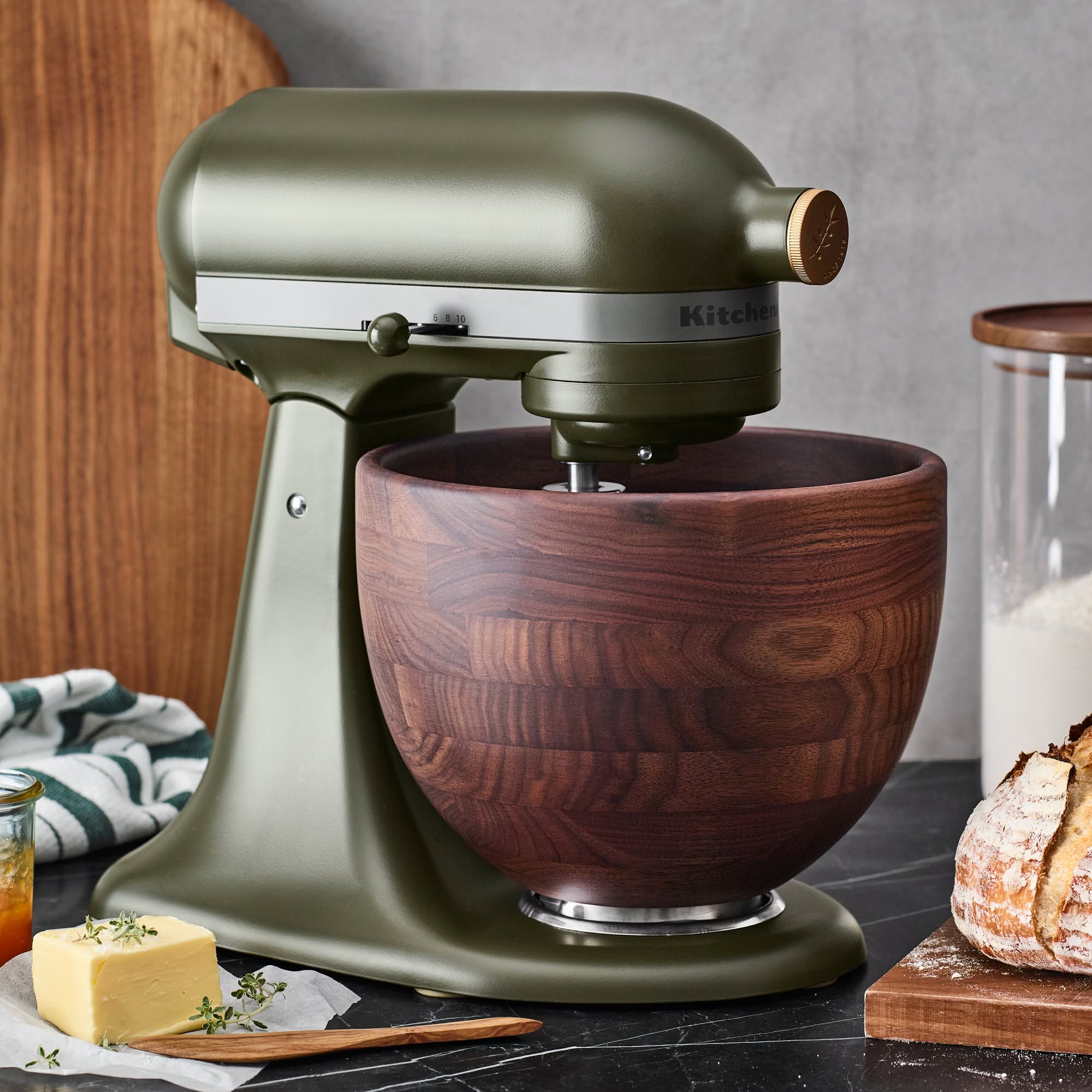 Olive green stand mixer with a dark wood mixing bowl on a kitchen counter, next to a stick of butter.
