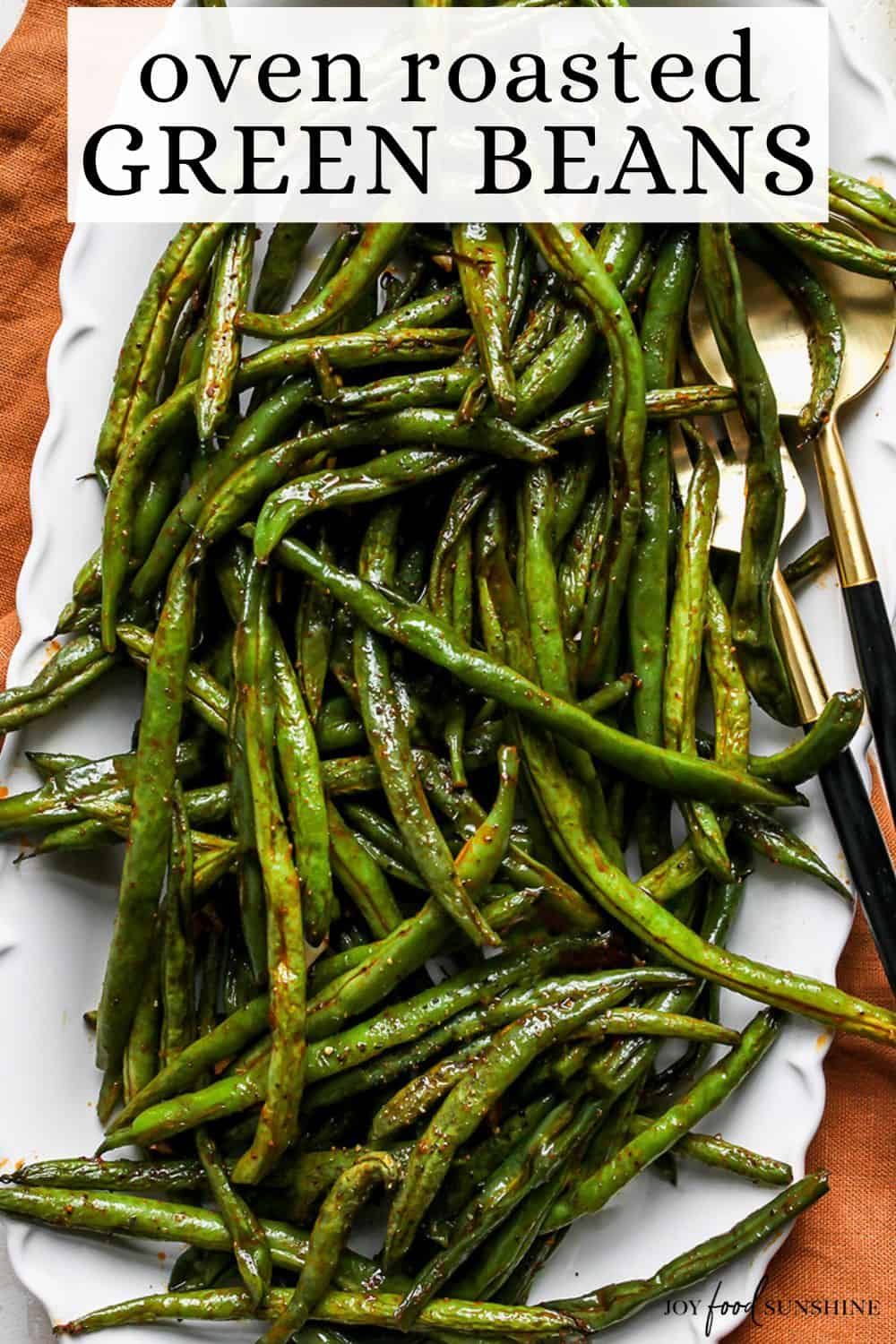 Close-up of a serving platter filled with glistening oven-roasted green beans, with two gold-handled serving utensils.