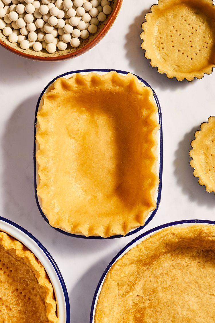 Several raw and partially baked pie crusts in dishes and mini tart molds, with a bowl of pie weights nearby.