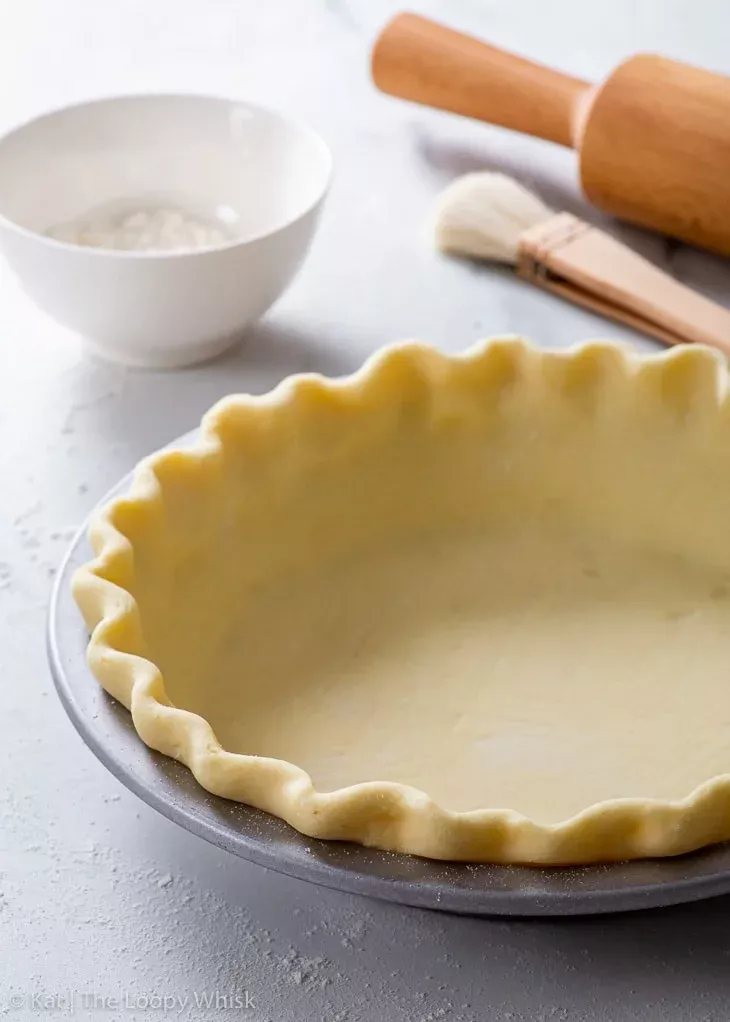 Unbaked pie crust with fluted edges inside a metal pie dish, with a small bowl and rolling pin in the background.