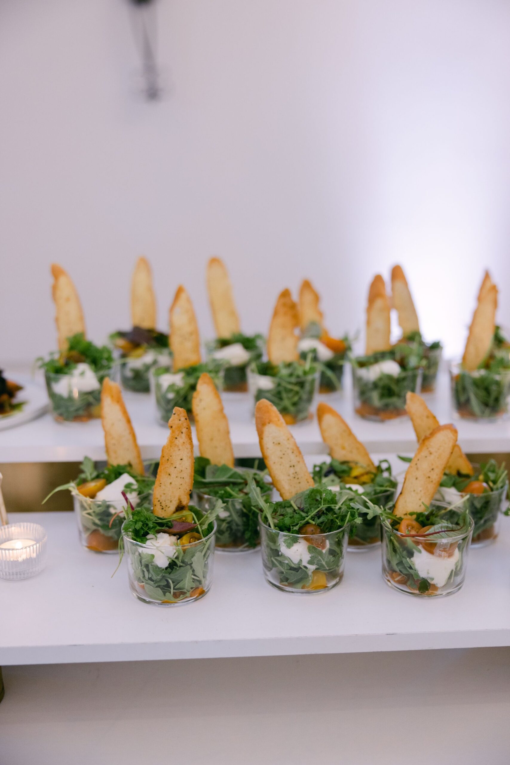 Many individual servings of caprese salad, layered with greens, cheese, and tomatoes, topped with toasted bread, arranged on…