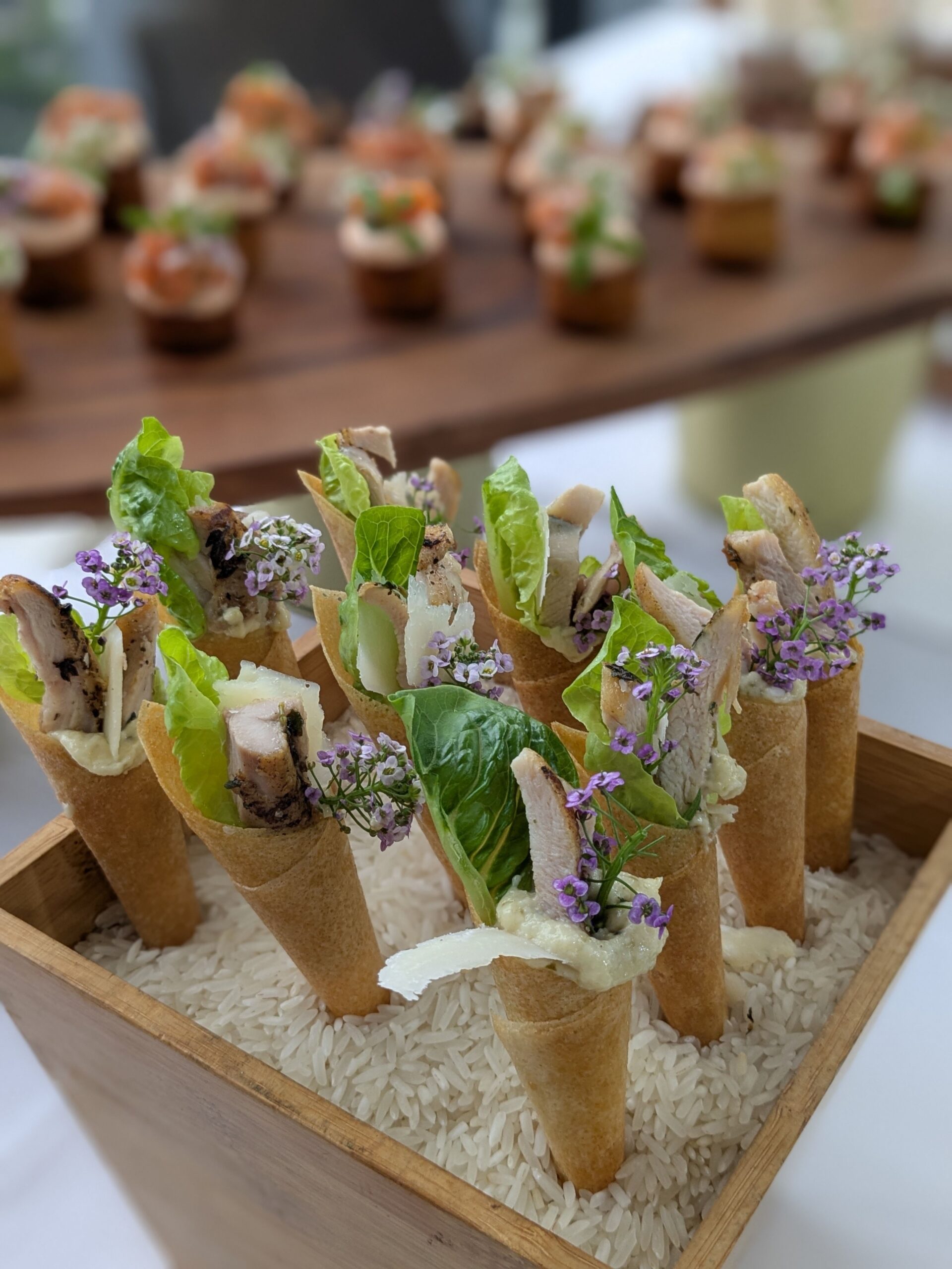 Savory cones filled with chicken, lettuce, sauce, and cheese, garnished with small purple flowers, displayed in a wooden box…
