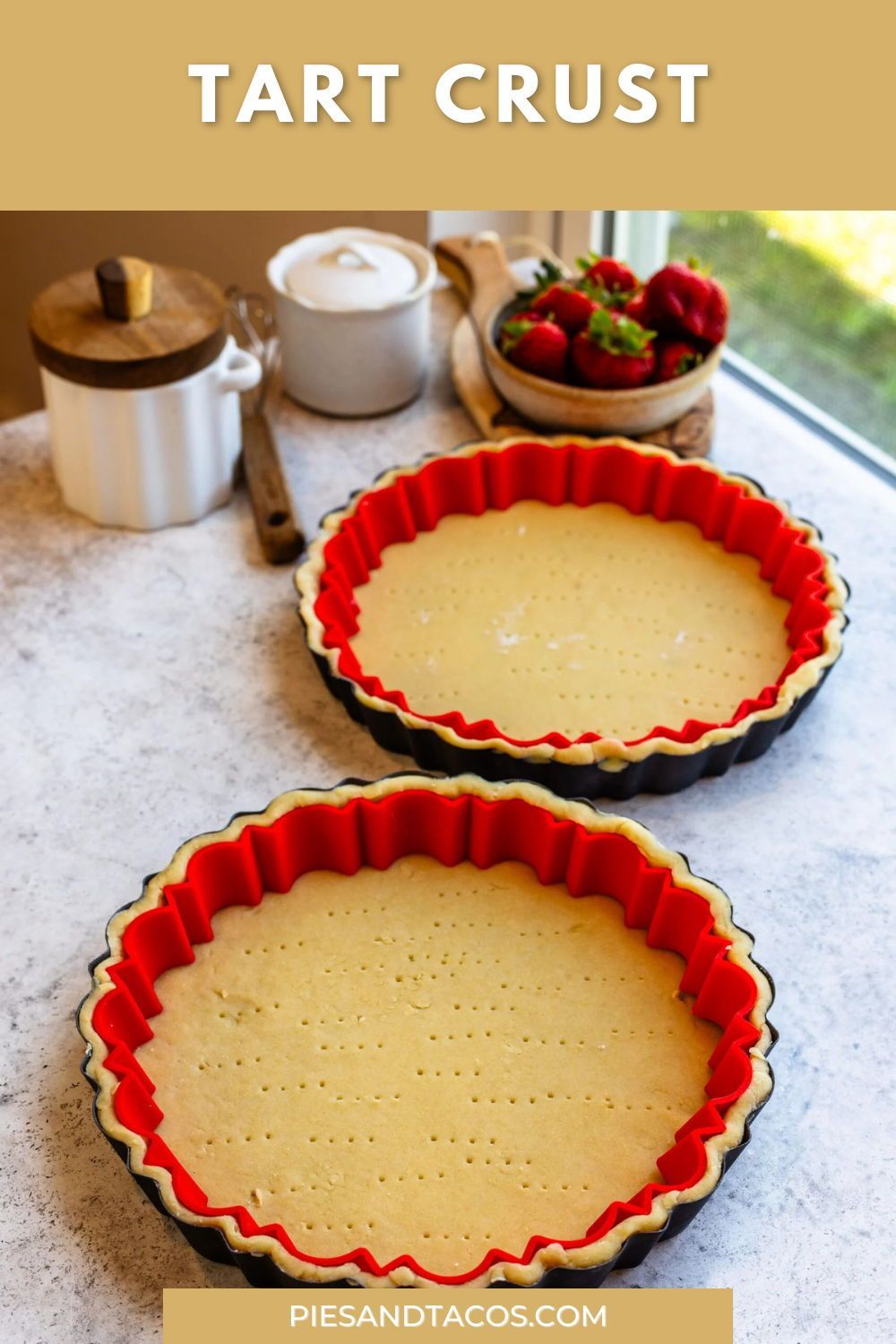 Two unbaked tart crusts in fluted metal tart pans with red silicone inserts, on a counter next to fresh strawberries.
