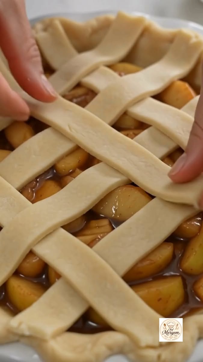Hands weaving strips of pie dough into a lattice pattern over a pie dish filled with sliced apples.