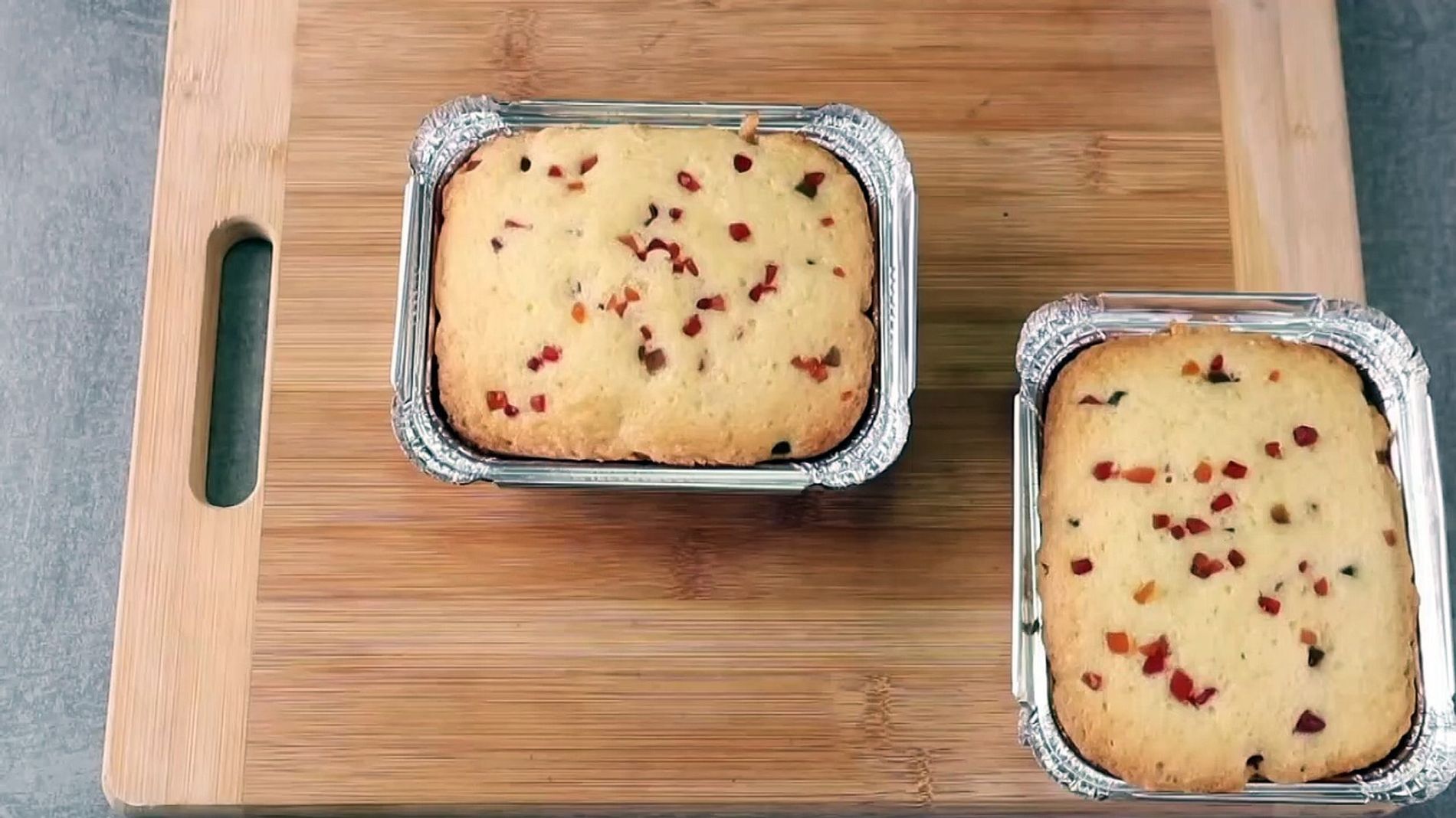 Two small rectangular cakes baked in disposable aluminum foil pans, topped with candied fruit bits, resting on a wooden…