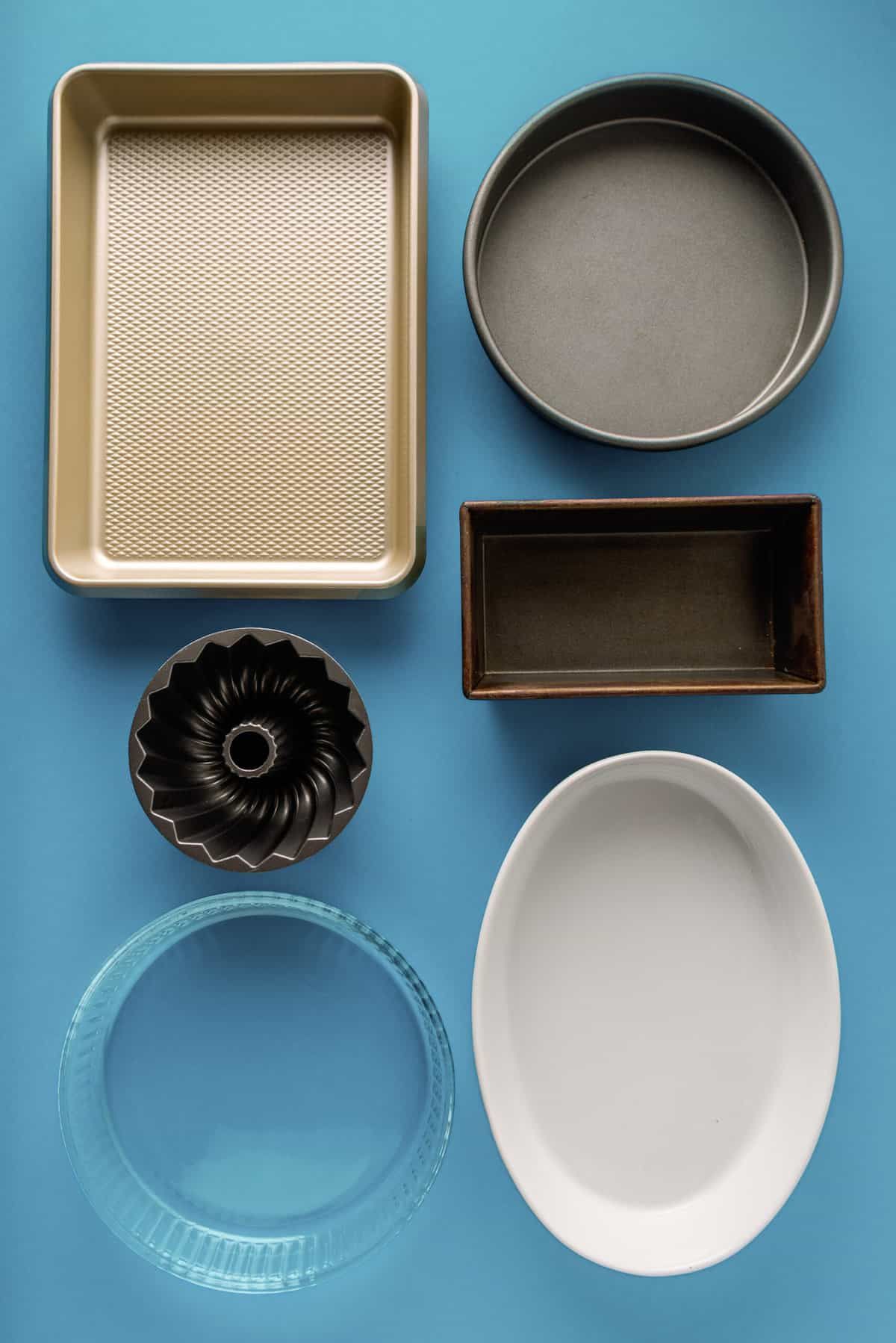 Rectangular baking pan, round cake pan, loaf pan, bundt pan, glass pie dish, and white oval casserole dish arranged on a…
