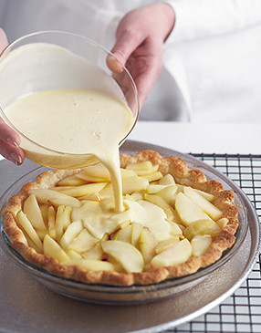 Hands pouring a creamy custard mixture over sliced apples in a pie crust set in a glass pie dish.