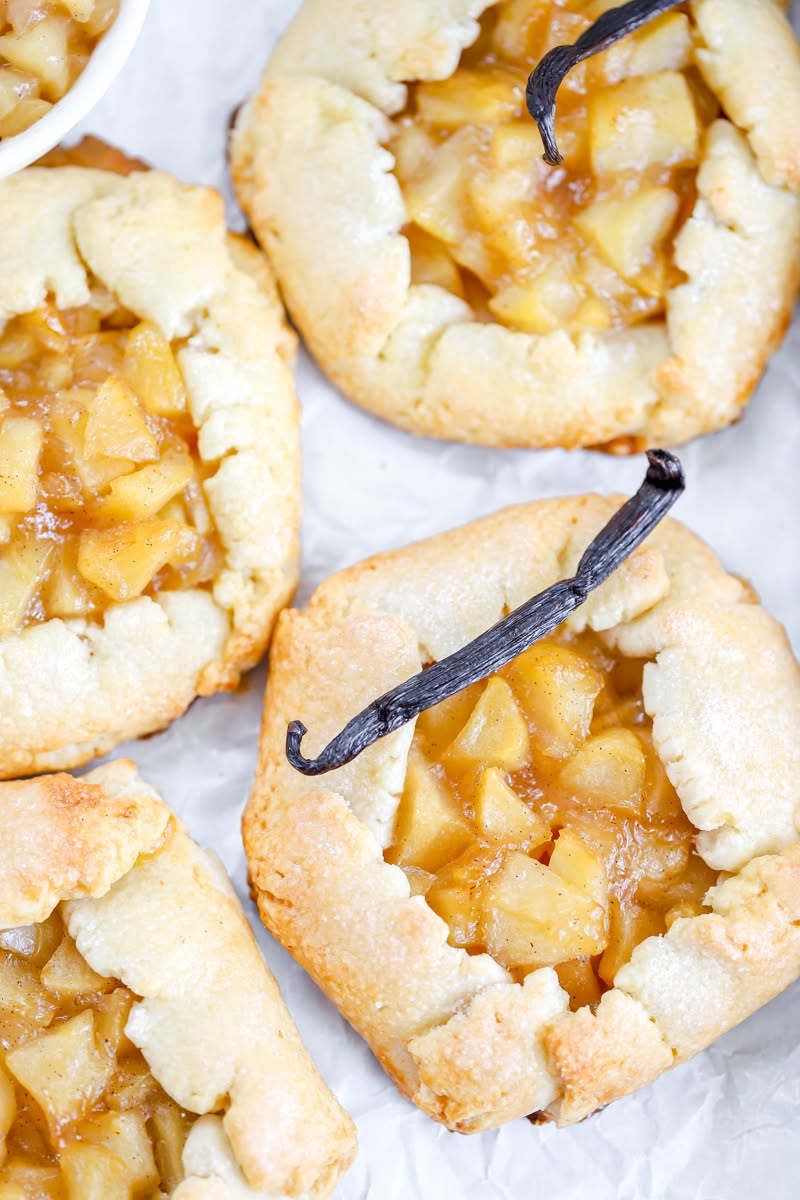 Several mini apple galettes with flaky crusts and chunky apple filling. Two have a whole vanilla bean laid on top.