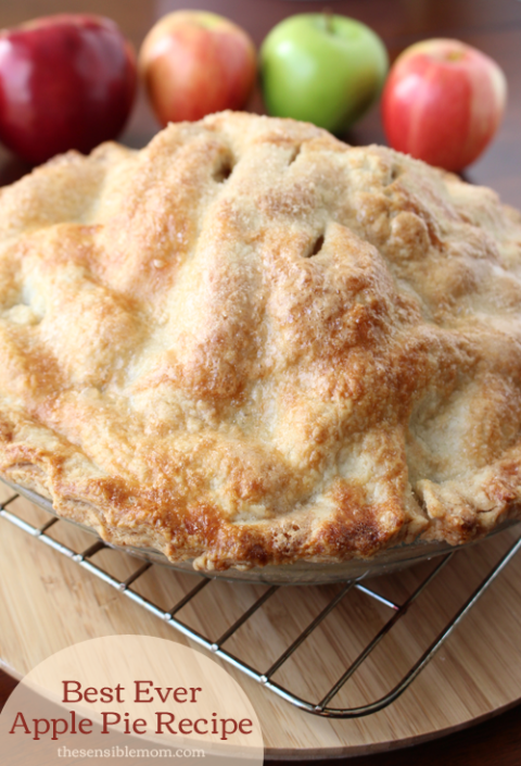 Baked apple pie on a cooling rack, with whole red and green apples in the background.
