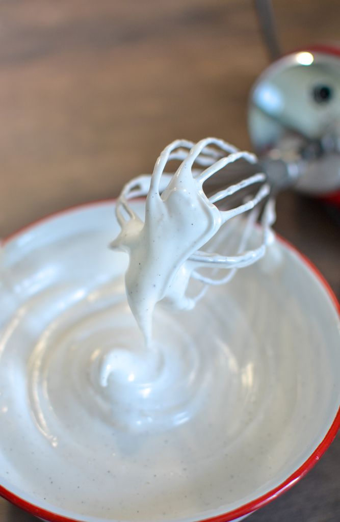 Close-up of a stand mixer whisk covered in stiff, white aquafaba meringue mixture with tiny black vanilla bean flecks, held…