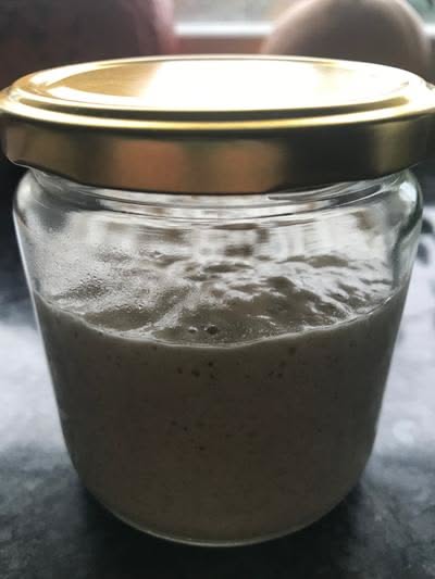 Small glass jar with a gold lid containing a beige, bubbly sourdough starter