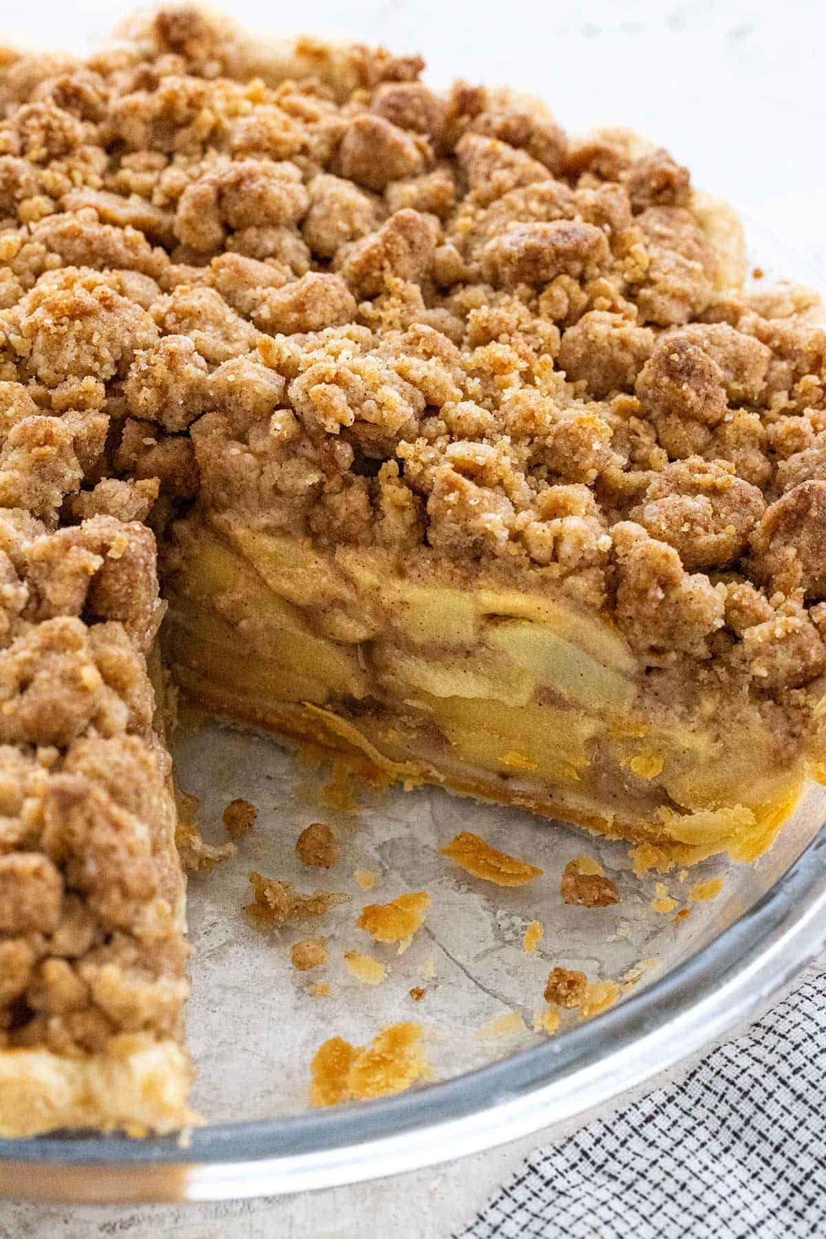 Close-up of a Dutch apple pie with a slice removed, showing thick layers of apple filling and a golden crumb topping.