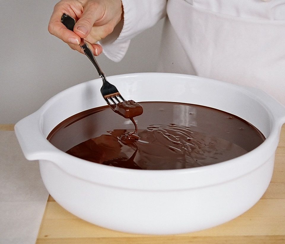 A hand holding a fork with a small, chocolate-dipped item over a bowl of melted chocolate.