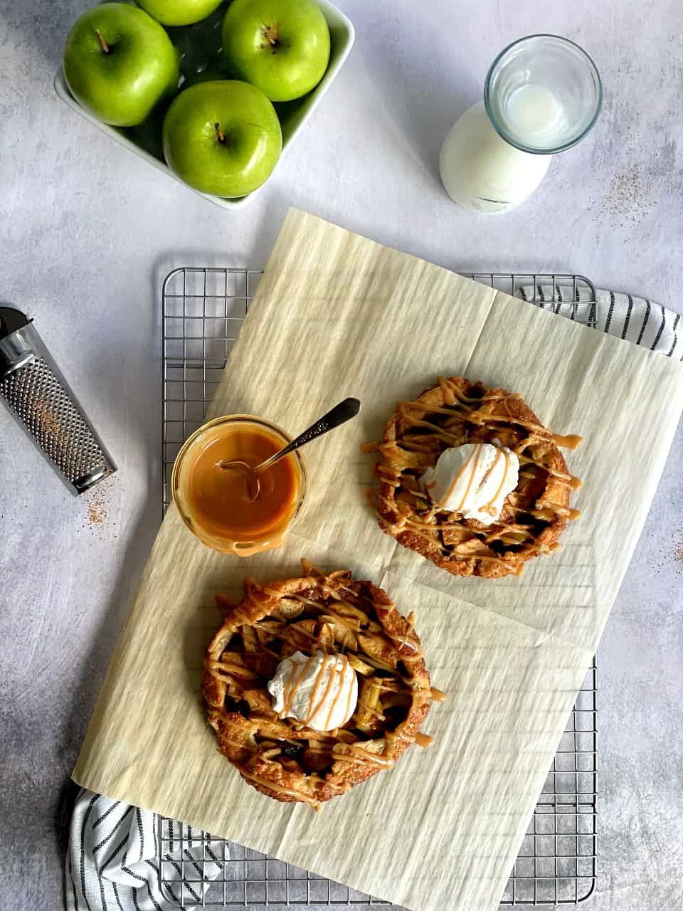 Two individual apple galettes with scoops of ice cream and caramel drizzle on parchment paper, next to a bowl of caramel…