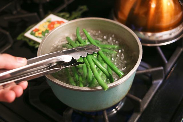 Tongs lift green beans from a turquoise pot of boiling water on a gas stove