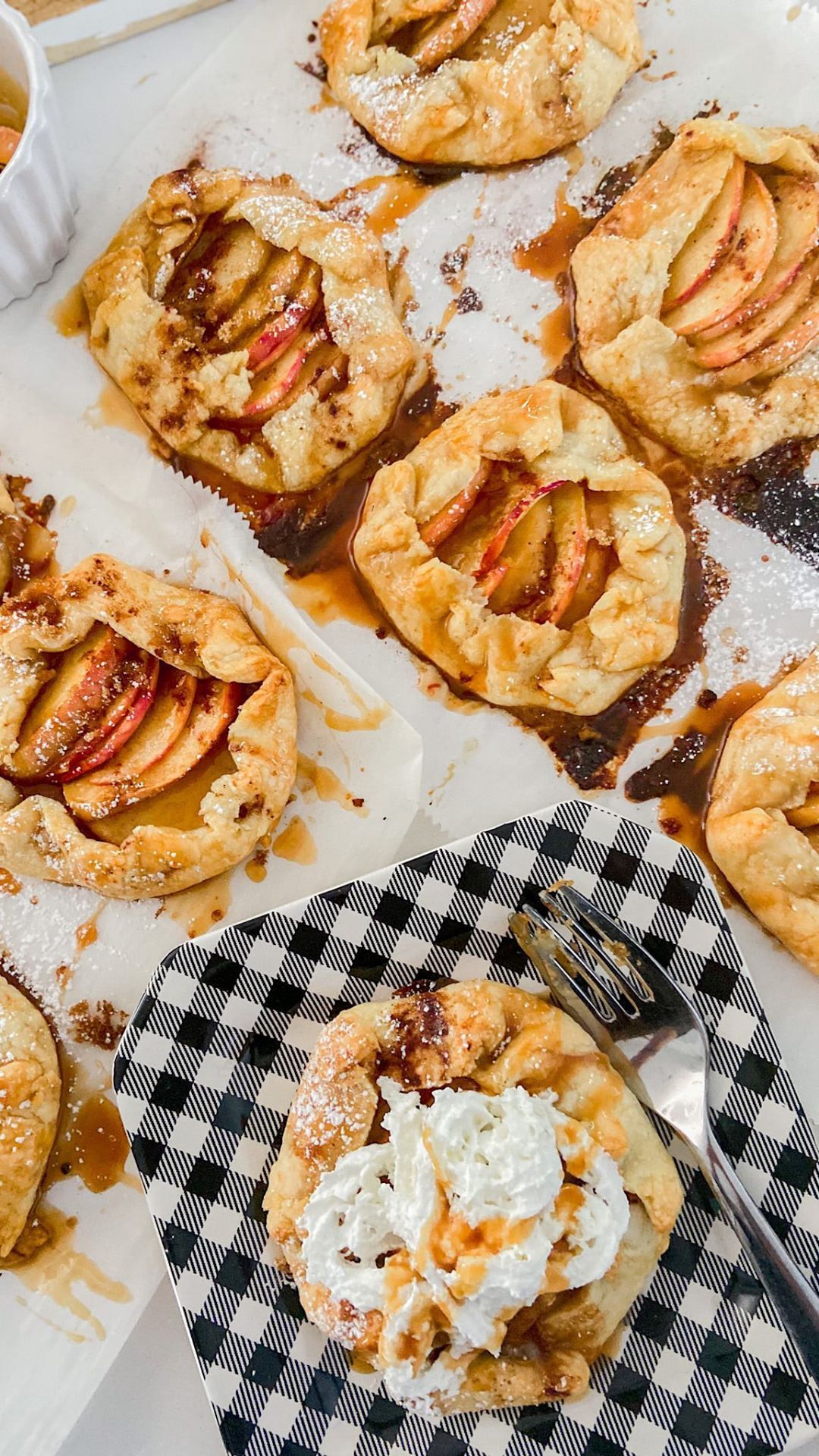 Several baked mini apple galettes on parchment paper, one on a black and white checkered plate with whipped cream and…