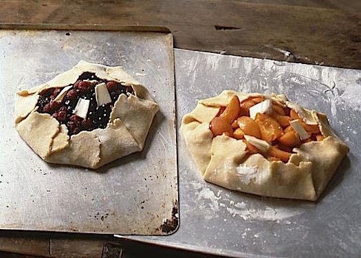 Two fruit galettes, one berry and one peach, resting on metal baking sheets, ready for baking.