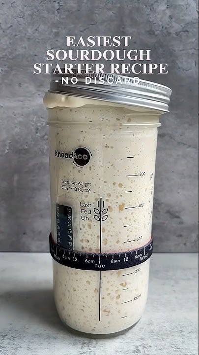 A glass jar contains an active sourdough starter, with a feeding schedule and temperature gauge wrapped around it.