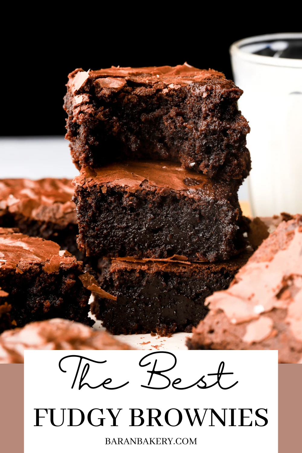A stack of three square fudgy brownies with crinkled tops, surrounded by other brownies, with a glass of milk in the…
