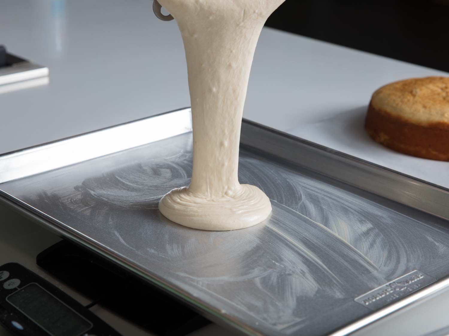 Thick cake batter being poured onto a sheet pan sitting on a digital scale, with a baked cake in the background.