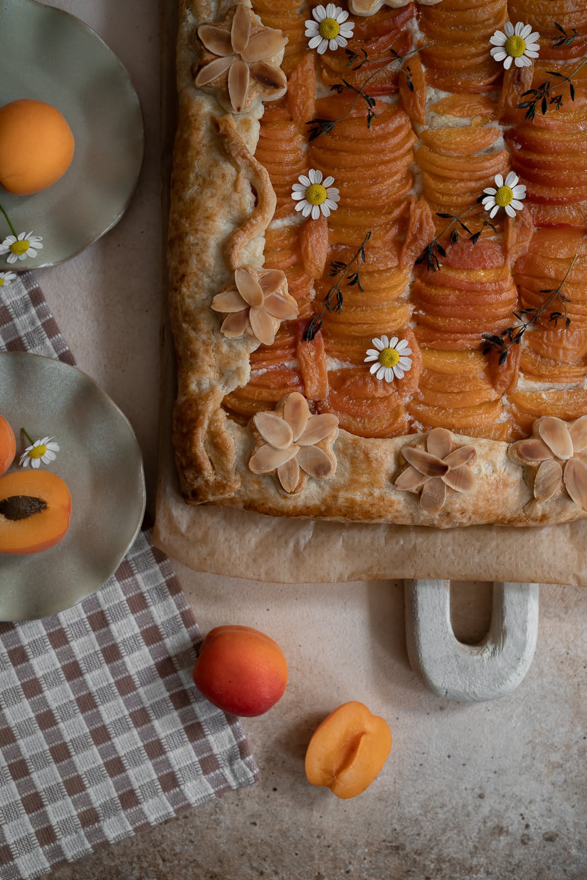 Close-up of a rectangular apricot galette with decorative almond slivers and small white flowers on top, next to whole and…