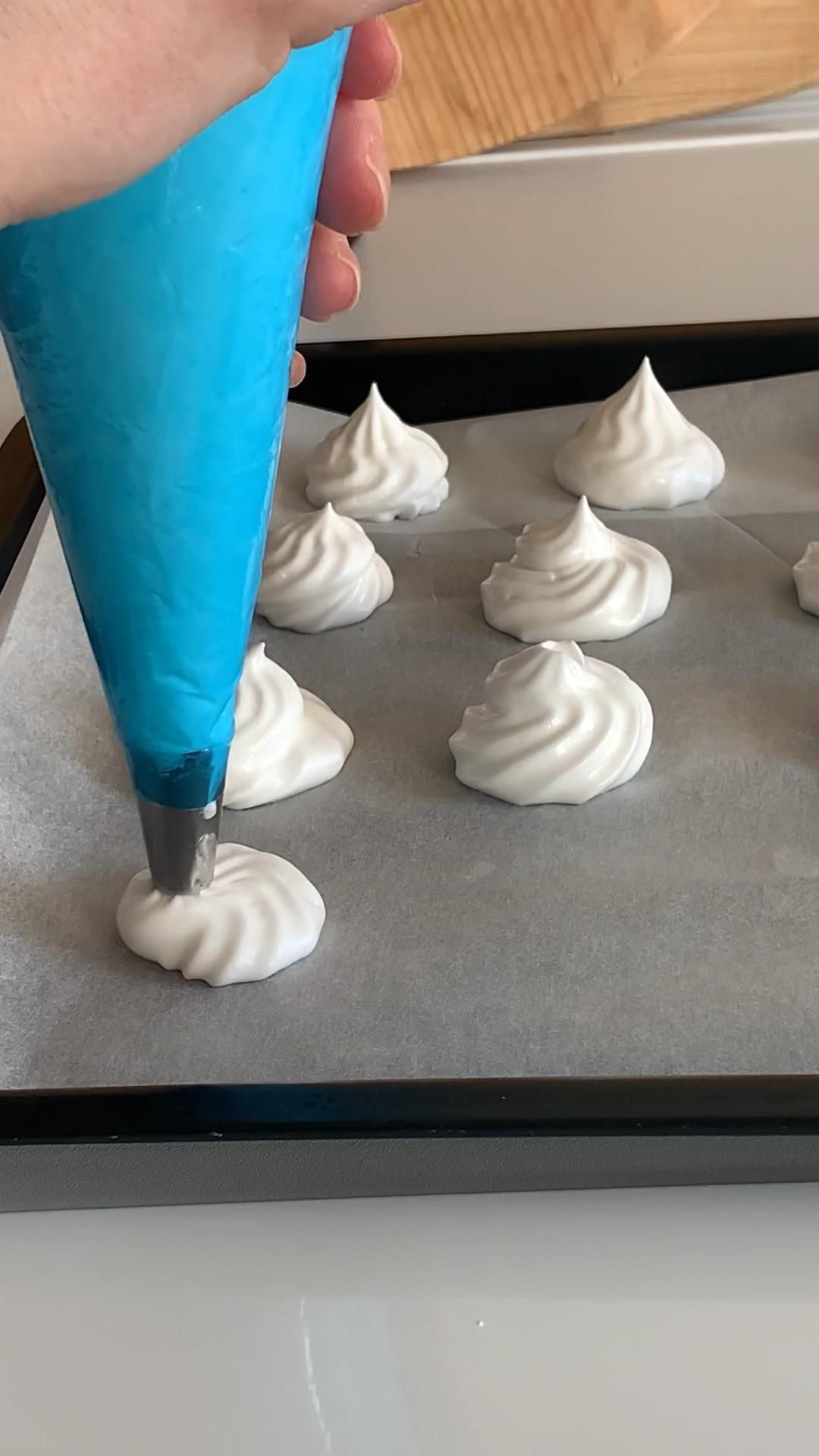 A hand squeezes a blue piping bag, forming white meringue swirls onto a parchment-lined baking sheet.