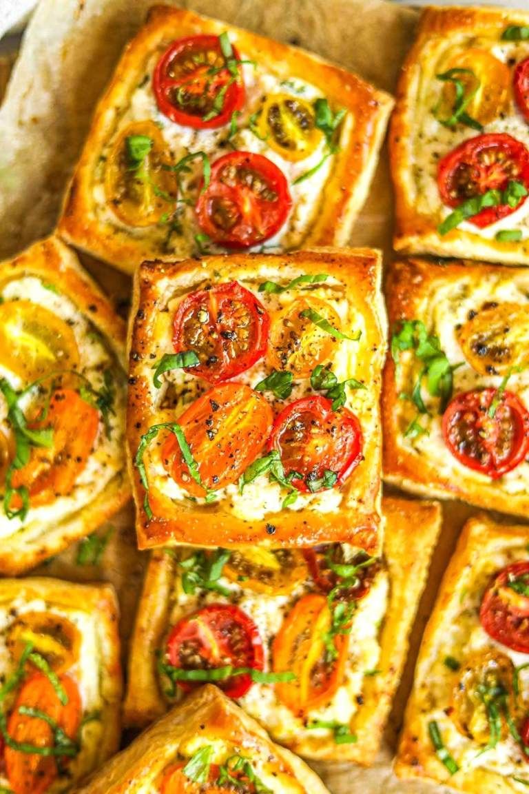 Close-up of baked rectangular puff pastry tarts with creamy filling, topped with sliced red and yellow cherry tomatoes and…